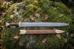 Damascus steel short Viking sword with brass cap pommel laid above its tan leather scabbard with brass throat and chape on a mossy rock
