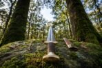 Damascus steel short Viking sword with brass guard and cord wrapped grip standing point up on a mossy boulder beside its scabbard