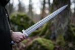 Damascus steel sword with a rune engraved guard held out in one hand, the patterned blade catching light against mossy boulders