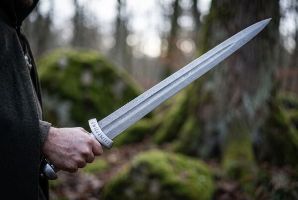 Damascus steel sword with a rune engraved guard held out in one hand, the patterned blade catching light against mossy boulders