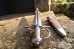 Damascus steel side sword with twin ring guards, curved knuckle bow and steel pommel, laid on a stone ledge beside its brown leather scabbard