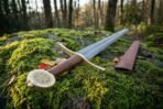 Damascus steel sword with pale cross guard, tan leather grip and Vegvisir disc pommel on a mossy rock beside a brown leather scabbard
