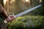 Damascus steel sword with Vegvisir pommel held level in a hand wearing a leather bracer against a soft green forest
