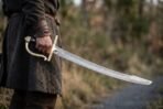 Damascus steel szabla sabre held point down at the side of a man in a wool tunic, brass hilt catching the light