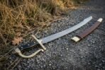 Damascus steel szabla sabre with black grip and rune cut brass knuckle bow lying on gravel beside its brown leather scabbard