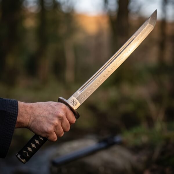 Damascus steel tanto sword with a fuller and black cord wrapped handle held up against a blurred autumn forest