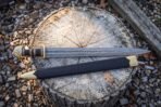 Damascus steel Viking sword with brass pommel and black grip beside a black leather scabbard on a weathered tree stump