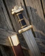Damascus steel Viking sword with rune engraved brass guard and black grip, close up of the fuller against an old fence