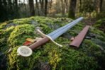 Damascus sword and brown leather scabbard resting on a moss covered boulder in a forest, low angle view toward the trees