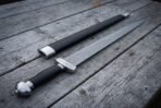 Damascus sword with a black grip and steel pommel laid on weathered planks, black scabbard with a steel throat placed beside it