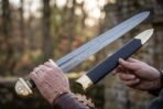 Damascus Viking sword drawn from a black leather scabbard with brass chape and held in two hands outdoors