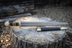 Damascus Viking sword with dark etched layered steel blade and black leather scabbard resting on a weathered tree stump in a wooded clearing