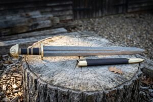 Damascus Viking sword with dark etched layered steel blade and black leather scabbard resting on a weathered tree stump in a wooded clearing