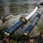 Damascus wakizashi with engraved brass tsuba and navy cord handle beside its black saya on a mossy river rock