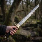 Dao sword with a curved patterned steel blade held out in one hand by its black wrapped grip, brass collar showing, woodland behind
