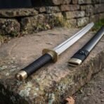 Close up of a dao sword hilt with a brass guard, black wrapped grip and brass pommel cap, black scabbard with brass throat beside it