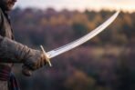 Deeply curved steel shamshir with brass cross quillons raised in a gloved hand against a misty autumn valley at dusk