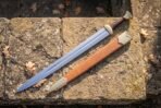 Double edged longsword with brass guard and pommel and cord wrapped grip lying beside its tan leather scabbard on sandstone