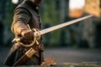Dueling sword with brass swept hilt held at arm's length by a man in a leather doublet with scabbard at his hip