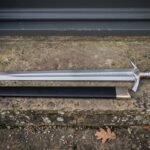 Elven sword replica with an engraved double edged blade and black leather scabbard resting on a weathered stone doorstep outside a stone cottage