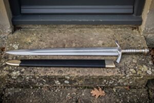 Elven sword replica with an engraved double edged blade and black leather scabbard resting on a weathered stone doorstep outside a stone cottage