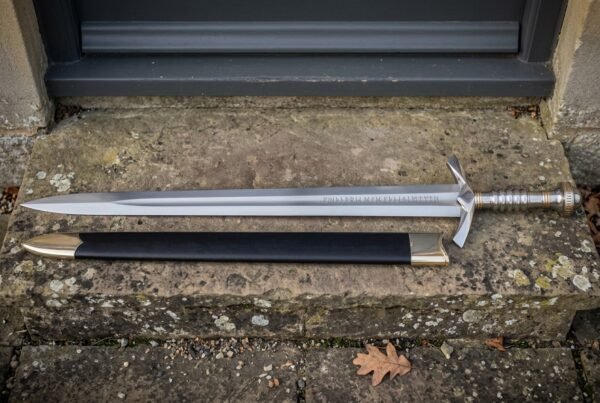 Elven sword replica with an engraved double edged blade and black leather scabbard resting on a weathered stone doorstep outside a stone cottage
