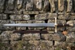 Fantasy longsword and dark brown scabbard resting on a low drystone wall, showing the etched fuller inscription and ribbed leather wrapped grip