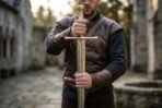 Fantasy longsword with brass toned cross guard and cream wrapped grip held upright point down by a man in a leather vest