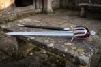 Full length basket hilt side sword with fullered blade laid on an old stone bench beside a black leather scabbard