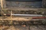 Full length dueling sword with brass hilt and polished steel blade on a stone step beside a russet leather scabbard