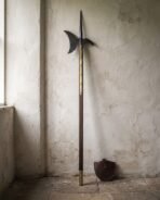 Full length halberd polearm standing upright against a plaster wall, crescent blade at the top and leather cover at its foot