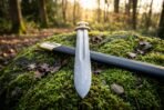 Fullered Damascus Norse sword blade crossed over its black scabbard with brass throat on a mossy boulder