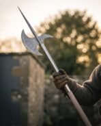 Gloved hand gripping the ash pole of a Damascus halberd with spike and crescent blade against a soft evening sky