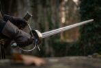 Gloved hand holding a Damascus rapier with steel cup hilt and knuckle bow, blade pointing across a dark courtyard