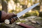 Gloved hand holding a Damascus steel arming sword by its dark leather grip, brass disc pommel and straight brass cross