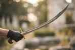 Gloved hand holding a steel shamshir sword with deep curve, brass cross guard and hooked wooden pommel outdoors