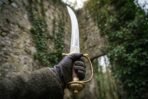 Gloved hand raising a curved talwar with polished blade, brass knuckle guard and disc pommel in an ivy covered ruin