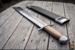 Grosse messer with pale wood grip, steel pins and pommel cap shown from the hilt end on weathered wood beside its black sheath