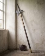 Halberd polearm with brass langets and dark walnut haft leaning in a window corner beside its round leather head cover