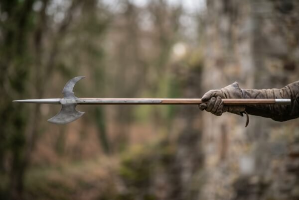Halberd with a crescent blade and spike held out level in a gloved hand against a blurred woodland backdrop
