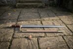 Hand forged 1095 high carbon steel Excalibur sword replica with gold tone guard, full length beside its tan scabbard on stone paving