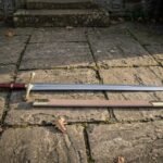 Hand forged 1095 high carbon steel Excalibur sword replica with gold tone guard, full length beside its tan scabbard on stone paving
