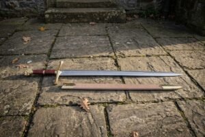 Hand forged 1095 high carbon steel Excalibur sword replica with gold tone guard, full length beside its tan scabbard on stone paving