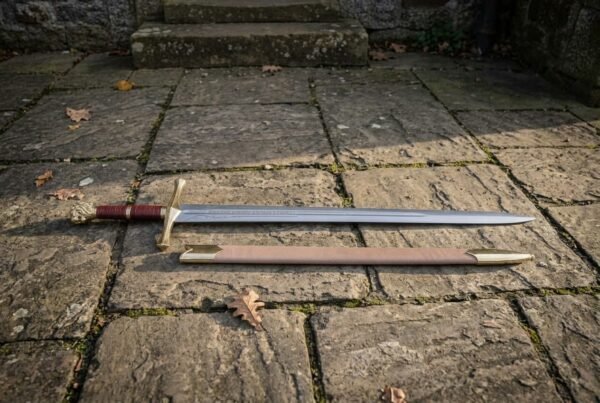 Hand forged 1095 high carbon steel Excalibur sword replica with gold tone guard, full length beside its tan scabbard on stone paving