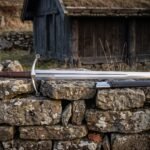 Hand forged arming sword with a polished fullered blade, leather grip and engraved valknut disc pommel resting in its scabbard on a drystone wall
