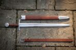Hand forged arming sword with scabbard and belt, polished fullered type XII blade and steel disc pommel, laid out on mossy grey flagstone paving.