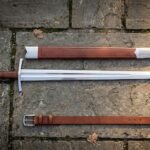 Hand forged arming sword with scabbard and belt, polished fullered type XII blade and steel disc pommel, laid out on mossy grey flagstone paving.