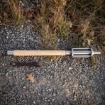 Hand forged bar mace with a fluted polished steel head and pale ash haft lying on a gravel path beside a brown leather wrist strap in low sun