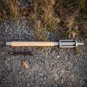 Hand forged bar mace with a fluted polished steel head and pale ash haft lying on a gravel path beside a brown leather wrist strap in low sun