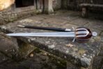 Hand forged basket hilt sword with a knotwork engraved steel shell guard and oxblood leather grip lying on a weathered stone garden bench beside its dark scabbard